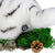 close-up of a white realistic tiger plush shown from a rearward angled perspective, featuring the 8 Billion Trees charm logo tag, placed on a patch of green moss with a small pinecone in front of it.  